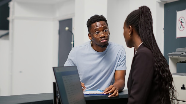 Paziente afroamericano che compila i documenti del referto medico alla reception dell'ospedale, parlando con l'addetto alla reception dell'appuntamento per la consultazione. Dipendente che fornisce assistenza all'uomo che scrive il modulo di controllo.