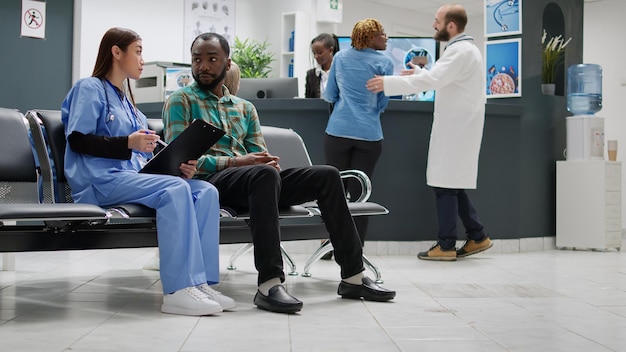 Paziente afroamericano e infermiera che parlano di assistenza sanitaria nella hall della sala d'attesa, facendo consulenza alla reception dell'ospedale. Diverse persone che fanno un esame di controllo presso la struttura. Scatto da treppiede.