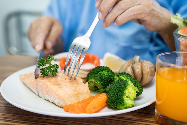 Paziente anziano asiatico della donna che mangia la prima colazione della bistecca di salmone con il cibo sano della verdura mentre si siede e ha fame sul letto in ospedale