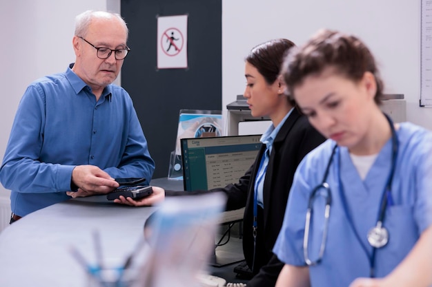 Paziente anziano in piedi al banco dell'ospedale che paga per il trattamento farmacologico con il telefono cellulare utilizzando il pagamento senza contatto. Persona anziana che ha visita di controllo consultazione con il medico