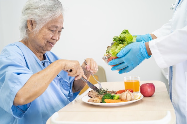 Paziente asiatico anziano o anziano della donna che mangia colazione e cibo sano vegetale con speranza e felice mentre è seduto e affamato sul letto in ospedale