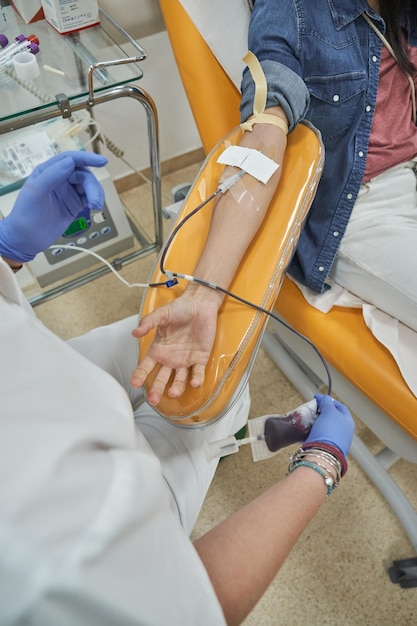 Paziente irriconoscibile in abiti casual seduta sulla poltrona medica e dona sangue vicino a una dottoressa professionista in un ospedale moderno
