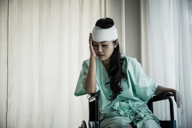 Pazienti femminili che hanno dolore nella sua testa dopo il ricovero in ospedale che si siede sulla sedia a rotelle nella stanza del paziente