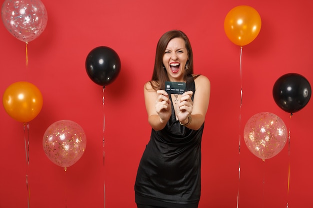 Pazza giovane donna in abitino nero che celebra, urla e mostra la carta di credito sulla fotocamera su mongolfiere sfondo rosso. San Valentino, felice anno nuovo, concetto di festa di compleanno mockup.