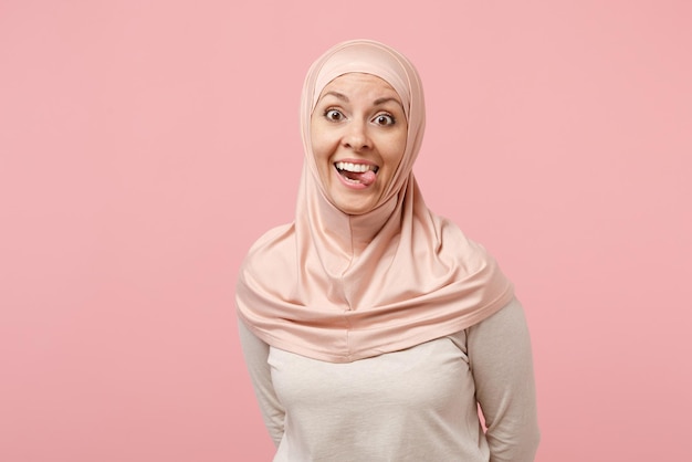 Pazzo divertente giovane donna musulmana araba in abiti leggeri hijab in posa isolata su sfondo rosa parete ritratto in studio. Concetto di stile di vita dell'Islam religioso della gente. Mock up spazio di copia. Mostrando la lingua.