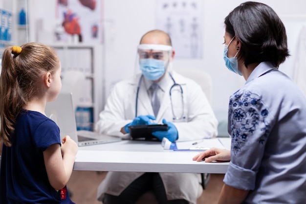 Pediatra che indossa una maschera facciale che tiene un tablet pc mentre consulta il bambino durante la pandemia clobal con coronavirus