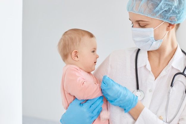 Pediatra medico femminile che indossa berretto medico, guanti e maschera protettiva, in piedi con la bambina in mano, esaminando il bambino in clinica, esprimendo emozioni positive.