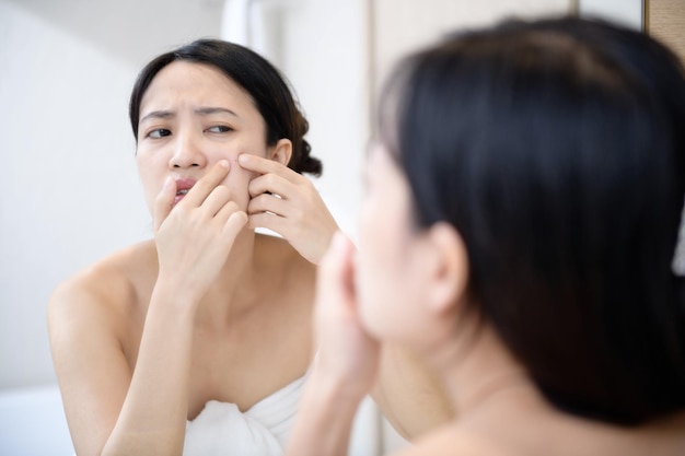 Pelle problematica Giovani donne asiatiche interessate che schioccano brufolo sulla guancia mentre si trovano vicino allo specchio in bagno giovani donne asiatiche con acne