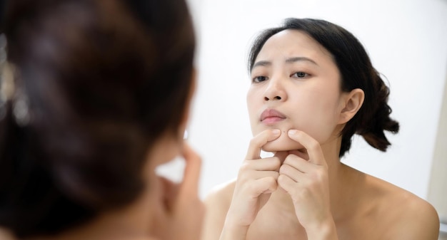 Pelle problematica Giovani donne asiatiche interessate che schioccano brufolo sulla guancia mentre si trovano vicino allo specchio in bagno giovani donne asiatiche con acne