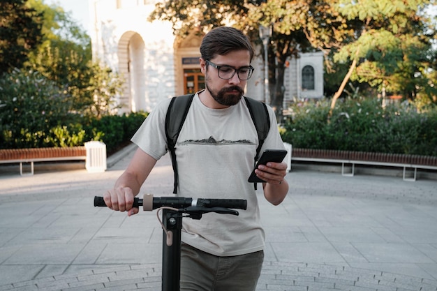 Pendolare uomo hipster con scooter elettrico all'aperto in città tramite smartphone