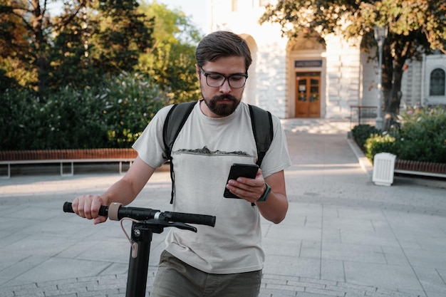 Pendolare uomo hipster con scooter elettrico all'aperto in città tramite smartphone