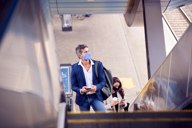Pendolari d'affari che guidano su una scala mobile alla stazione ferroviaria indossando maschere per il viso in DPI durante la pandemia