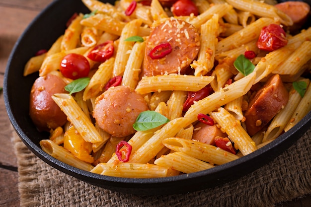 Penne al sugo di pomodoro con salsiccia, pomodori, basilico verde decorato in padella su un tavolo di legno