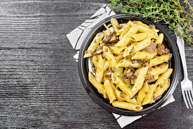 Penne con funghi selvatici in un piatto su un asciugamano da cucina, timo, forchetta e aglio su sfondo nero tavola di legno dall'alto