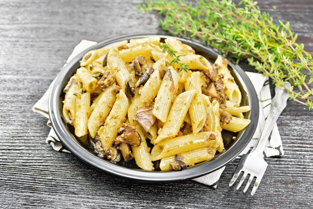 Penne con funghi selvatici in un piatto su un asciugamano, timo, forchetta e aglio su uno sfondo di tavola di legno