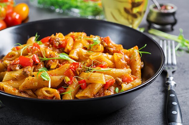 Penne in salsa di pomodoro con carne, pomodori decorati con germogli di pisello su un tavolo scuro.