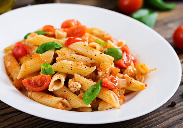 Penne in salsa di pomodoro con pollo, pomodori, decorato con basilico su un tavolo di legno. Cibo italiano. Pasta al ragù.