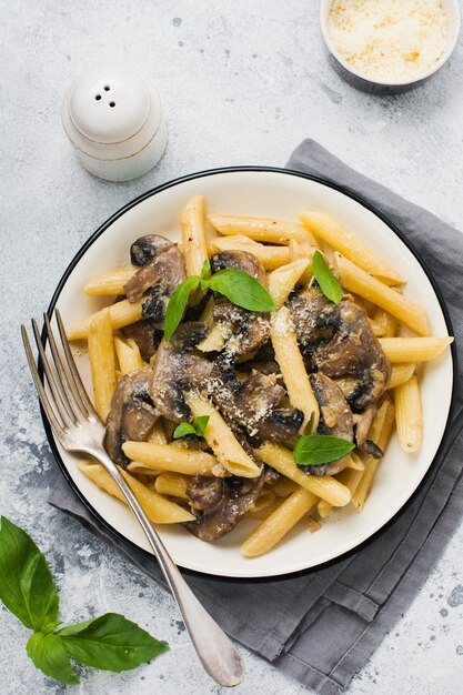 Penne rigate con funghi, parmigiano e foglie di basilico in un piatto di ceramica sul vecchio tavolo in cemento chiaro.