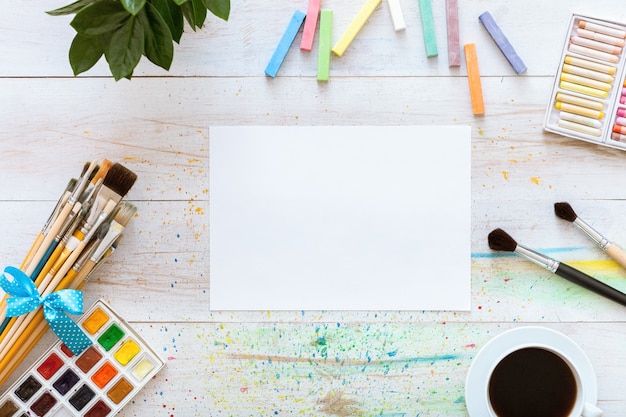 Pennelli, paintbox con acquerelli, pastelli, caffè e carta bianca su sfondo bianco in legno, fondale artistico, studio di artisti creativi, vista dall'alto dall'alto, piatto disteso con spazio di copia