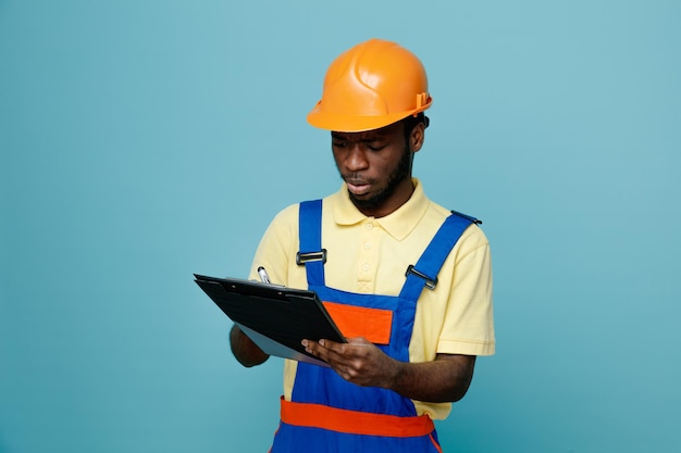 Pensando al giovane costruttore afroamericano in uniforme che tiene e scrivi negli appunti isolati su sfondo blu