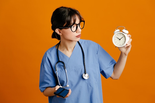 Pensando che tiene la sveglia parla al telefono giovane dottoressa che indossa uno stetoscopio fith uniforme isolato su sfondo arancione