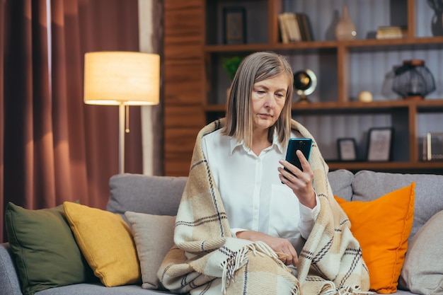 Pensando donna dai capelli grigi senior a casa seduta sul divano guardando lo schermo del telefono