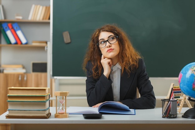 pensando guardando al lato giovane insegnante femminile che indossa occhiali seduto alla scrivania con strumenti scolastici in classe
