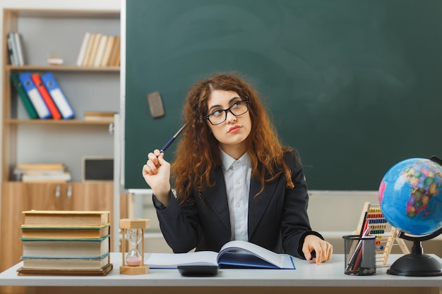 pensando guardando al lato giovane insegnante femminile che indossa occhiali tenendo il puntatore seduto alla scrivania con strumenti scolastici in classe