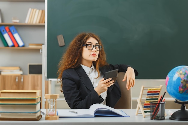 pensando guardando in alto giovane insegnante femminile che indossa occhiali tenendo la calcolatrice seduto alla scrivania con strumenti scolastici in classe