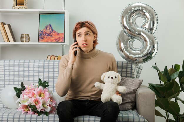 Pensare guardando il lato bello ragazzo durante la giornata delle donne felici che tiene l'orsacchiotto parla al telefono seduto sul divano nel soggiorno