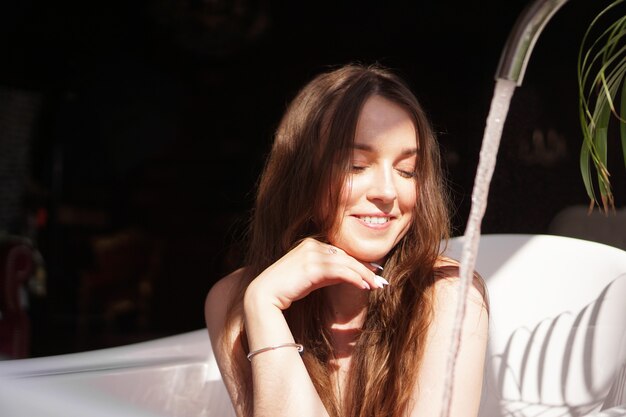 Pensare positivo. Ragazza affascinante in un bagno luminoso con una pianta verde. Una donna felice aspetta che la vasca da bagno si riempia d'acqua