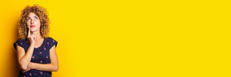 Foto pensierosa giovane donna riccia alzando lo sguardo tenendo il dito sul mento su uno sfondo giallo banner