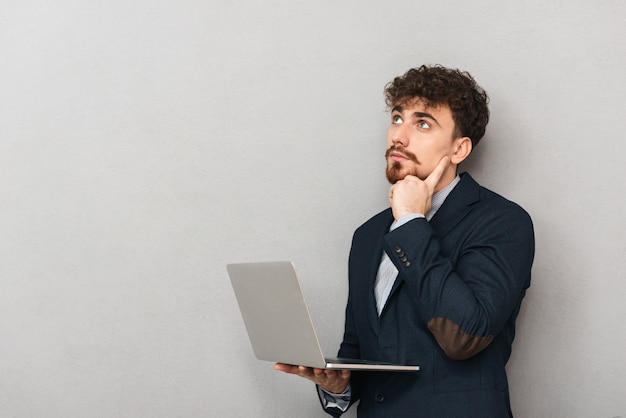 Pensieroso giovane imprenditore che indossa tuta in piedi isolato su grigio, lavorando su un computer portatile