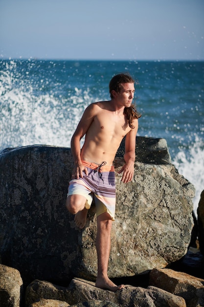 Pensieroso giovane senza camicia che si appoggia su una grande roccia sulla spiaggia del mare e distoglie lo sguardo