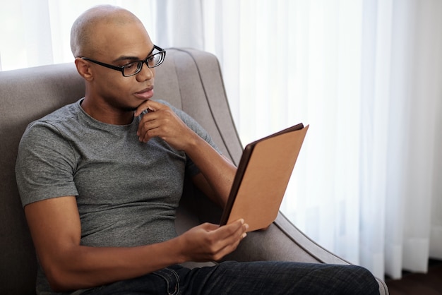 Pensieroso giovane uomo nero calvo con gli occhiali seduto in poltrona e leggere un libro interessante su tablet PC