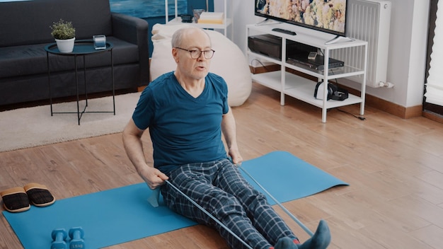 Pensionato che usa la fascia di resistenza per allungare i muscoli sulla stuoia di yoga. Persona anziana che tira la cintura elastica per fare esercizio fisico e attività di fitness. Allenamento uomo attivo per la flessibilità