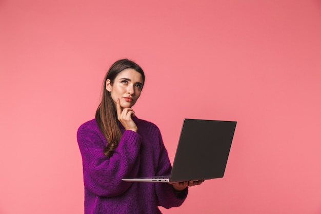 Pensive giovane ragazza che indossa un maglione in piedi isolato su rosa, tenendo il computer portatile