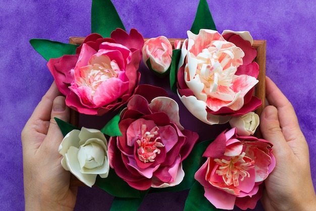 Peonie decorative fatte a mano in foamiran su pannello di legno che tiene le mani di una donna sullo sfondo viola