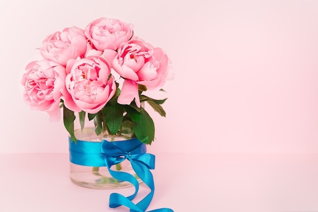 Peonie rosa delicate in un vaso di vetro con un nastro di raso blu. Elegante sfondo floreale rosa. Copia spazio. Biglietto d'auguri. Sfondo floreale minimalista