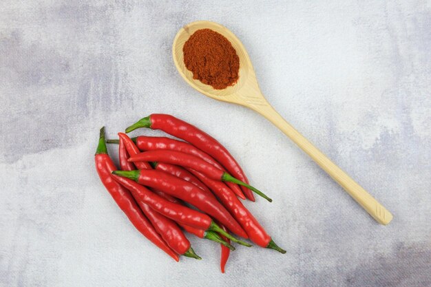 Peperoncini roventi macinati in un cucchiaio di legno e baccelli di peperoncino rosso su uno sfondo grigio astratto