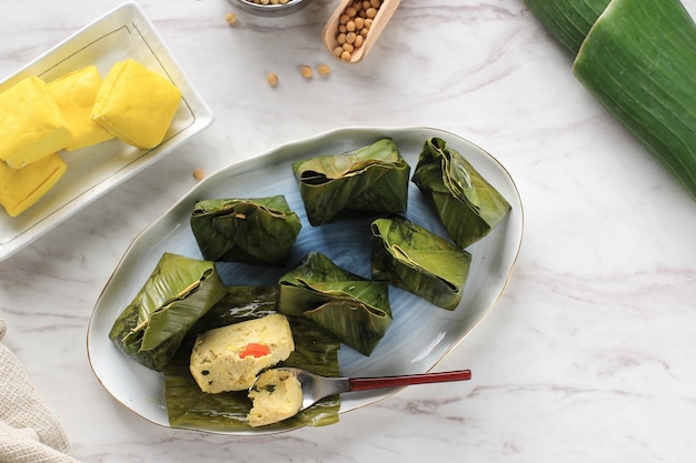 Pepes Tahu è il tofu speziato indonesiano avvolto con foglie di banana e cotto a vapore, cibo tipicamente indonesiano di Giava occidentale (sudanese). Tofu al vapore con basilico asiatico, sfondo bianco