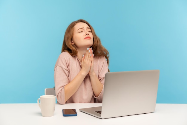 Per favore, dio aiuta la giovane donna dipendente seduta sul posto di lavoro e tenendo gli occhi chiusi pregando cordialmente supplicando con disperata espressione sincera indoor studio shot isolato su sfondo blu