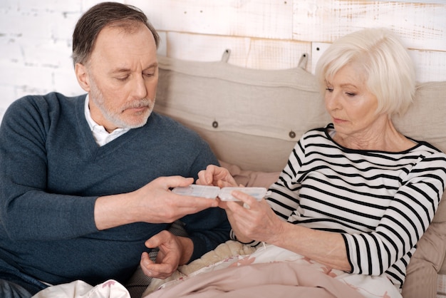 Per meglio. L'uomo anziano sta dando un caso di pillole alla sua anziana moglie ammalata sdraiata sul letto coperto di coperta calda.
