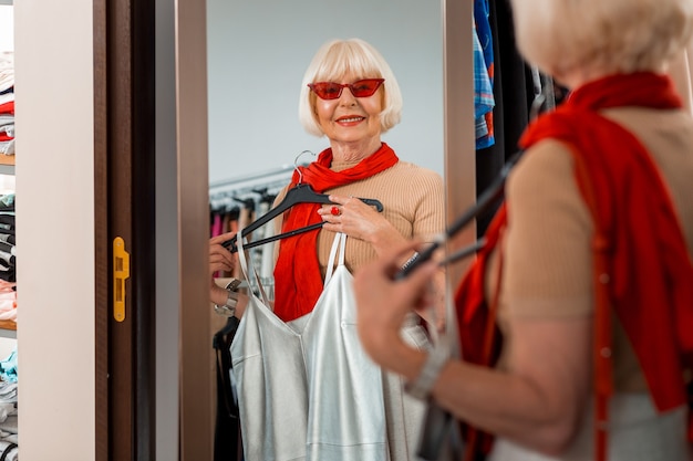 Per sempre giovani. Mezzo busto di donna anziana elegante che indossa occhiali da sole rossi e guardando nello specchio dello shopping con abito estivo in mano ammirando il suo riflesso