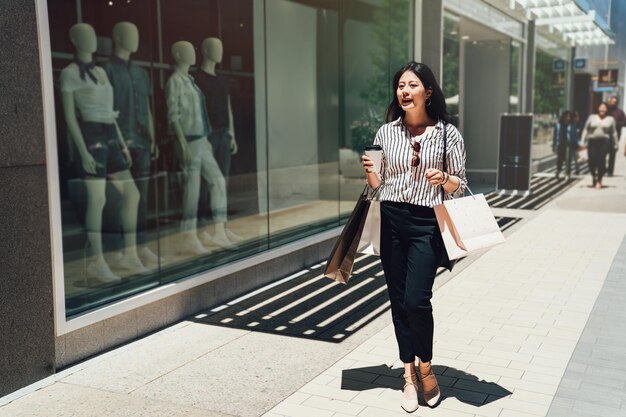 per tutta la lunghezza della bella giovane donna asiatica che trasporta la borsa della spesa. Ragazza carina che cammina su una strada cittadina dopo lo shopping in vendita estiva nel centro commerciale di Stanford. signora che tiene una tazza di caffè vicino alla finestra.