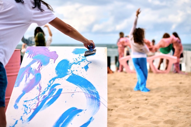 Performance artistica all'aperto con pittura a goccia con ragazze danzanti sulla spiaggia di mare sabbioso Artista pittore che disegna su tela bianca immagine astratta nella tecnica di arte astratta di pittura a goccia Festival di arte creativa