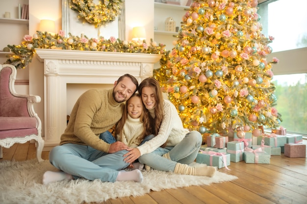 Periodo natalizio. Giovane famiglia felice seduta sul pavimento vicino all'albero di Natale e sentirsi a proprio agio