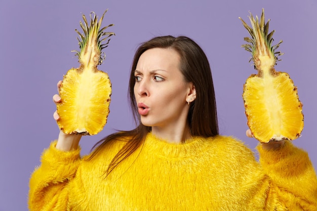 Perplessa giovane donna in maglione di pelliccia tenere in mano guardando metà di frutta fresca di ananas maturo isolato su sfondo viola pastello. Lo stile di vita vivido delle persone si rilassa il concetto di vacanza. Mock up spazio di copia.