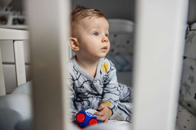 Perplesso affascinante bambino biondo seduto nella sua culla al mattino, tenendo in mano il suo giocattolo e guardando lontano.