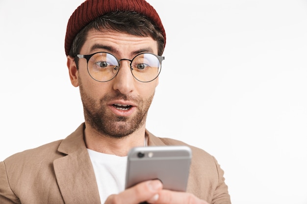 perplesso uomo che indossa un cappello hipster che sorride mentre tiene e utilizza lo smartphone isolato su un muro bianco